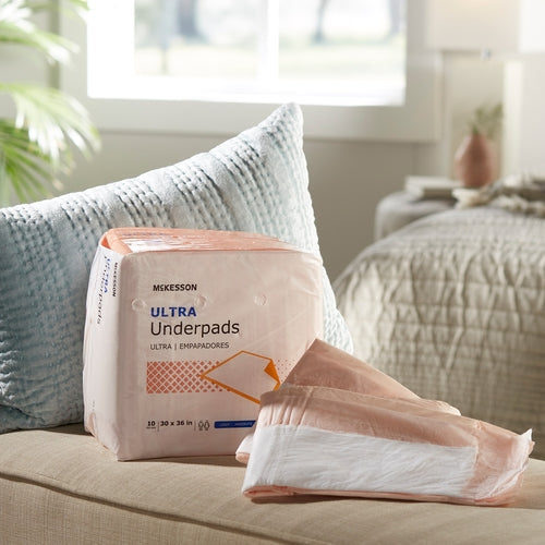 McKesson Ultra Underpads package with several underpads fanned out beside it, displayed on a beige sofa near a bed