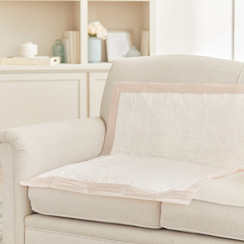 Disposable underpads laid flat on a cream upholstered sofa seat and back in a bright living room setting