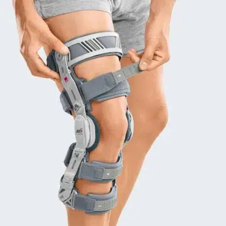 M.4s OA Comfort Compact Knee Brace worn on left leg, shown from front with person adjusting the gray rigid frame and straps