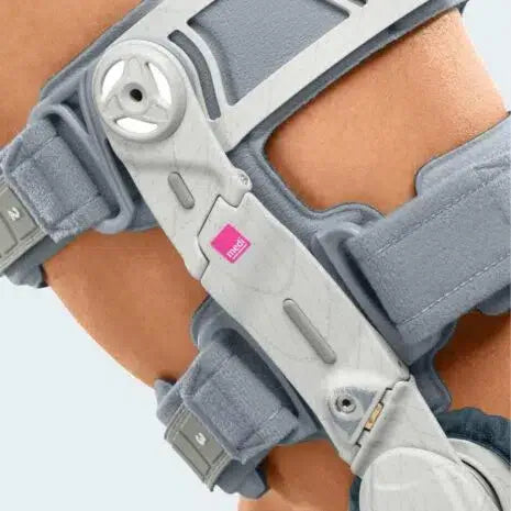 Knee brace with hinged metal joint, dial adjustment wheel, gray padded straps, and pink brand label worn on a leg