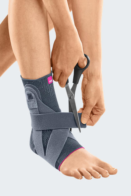 Levamed Active Ankle Support in gray with pink trim being adjusted on a foot using scissors to cut a strap tab