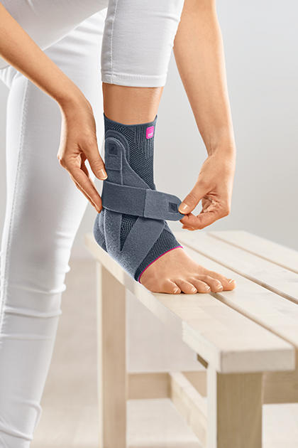 Levamed Active Ankle Support in gray with pink trim being fastened around a woman's ankle resting on a wooden bench