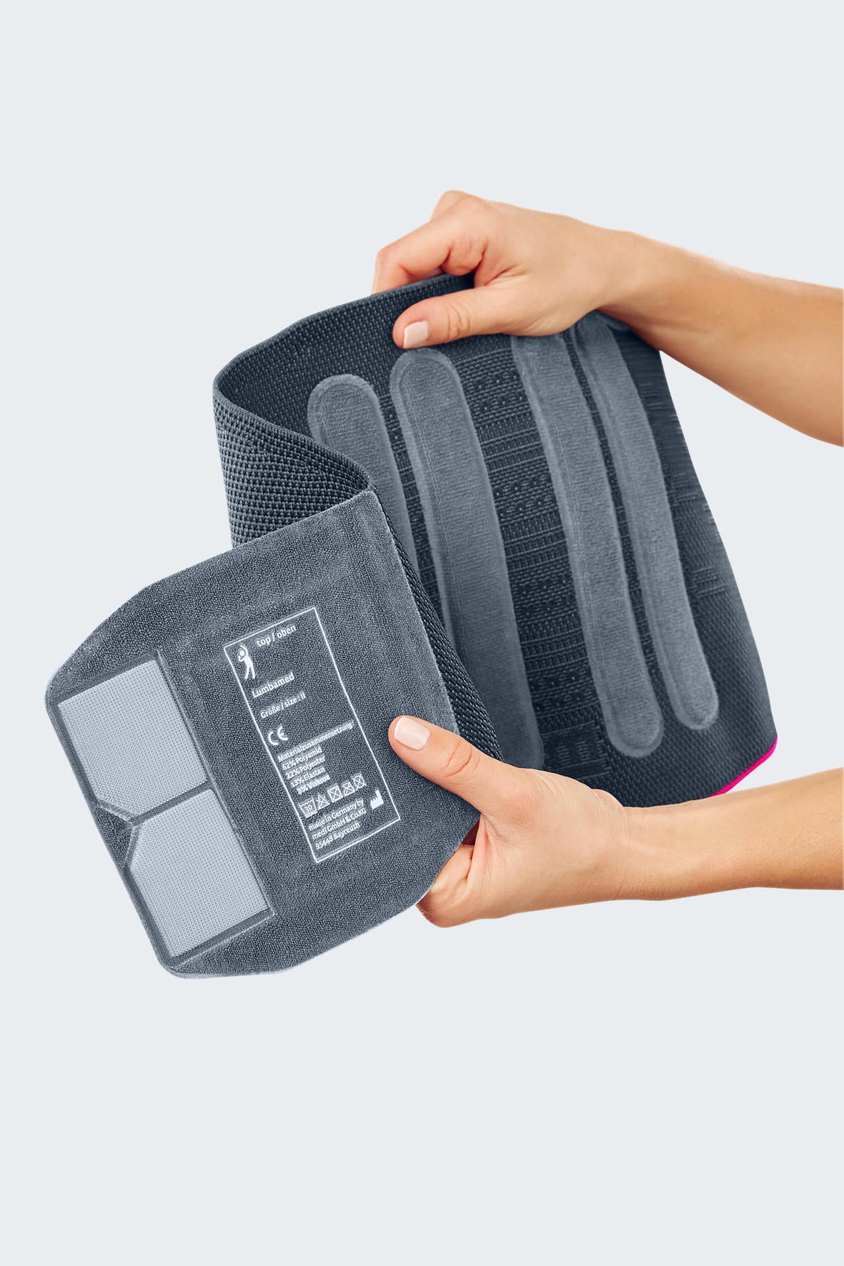 Lumbamed Basic lumbar support brace held open by two hands, showing interior stays and hook-and-loop closure, gray with pink