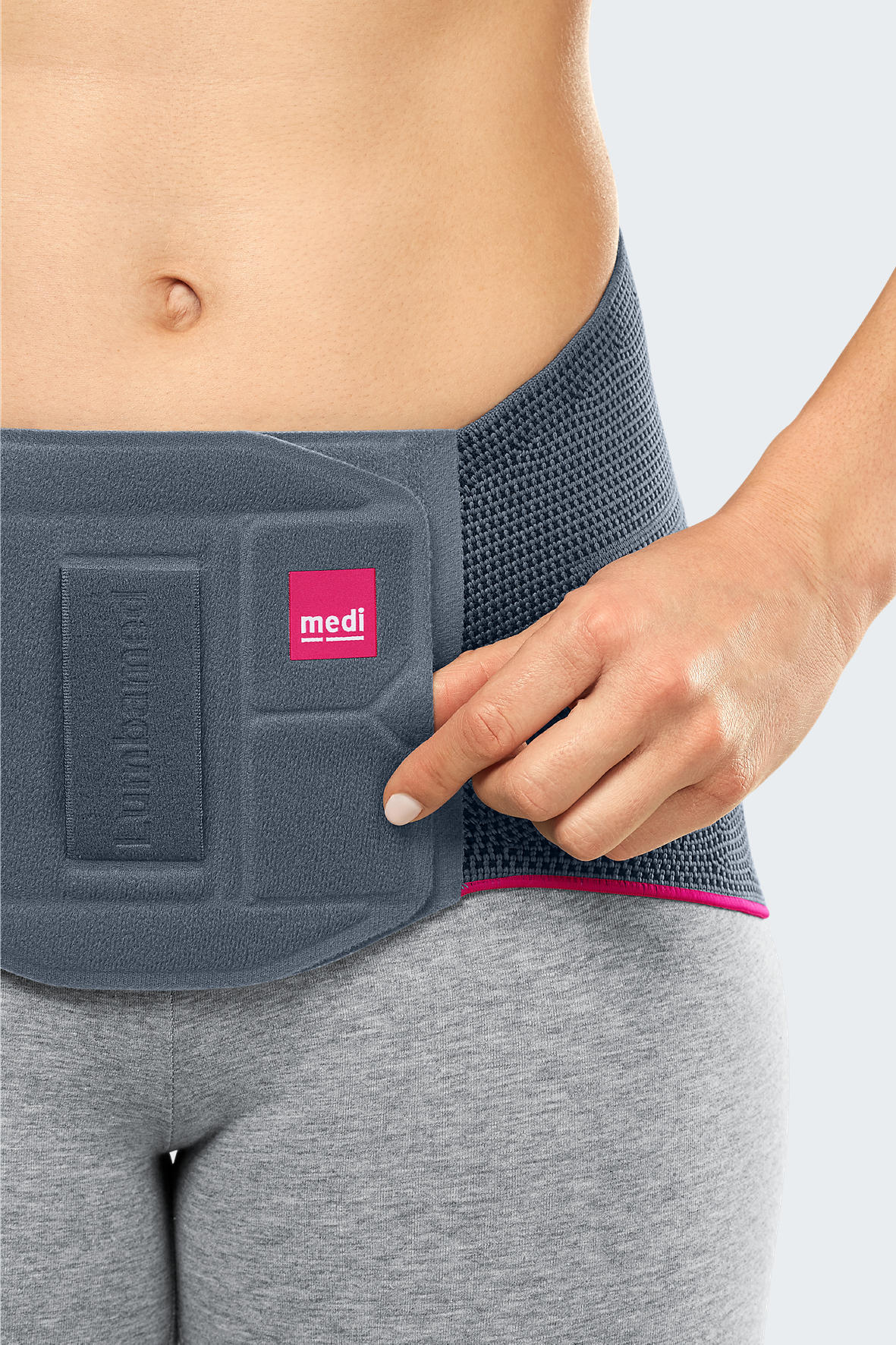 Lumbamed Basic lumbar support brace in gray with pink trim, worn by a woman, shown from front with hand adjusting fit