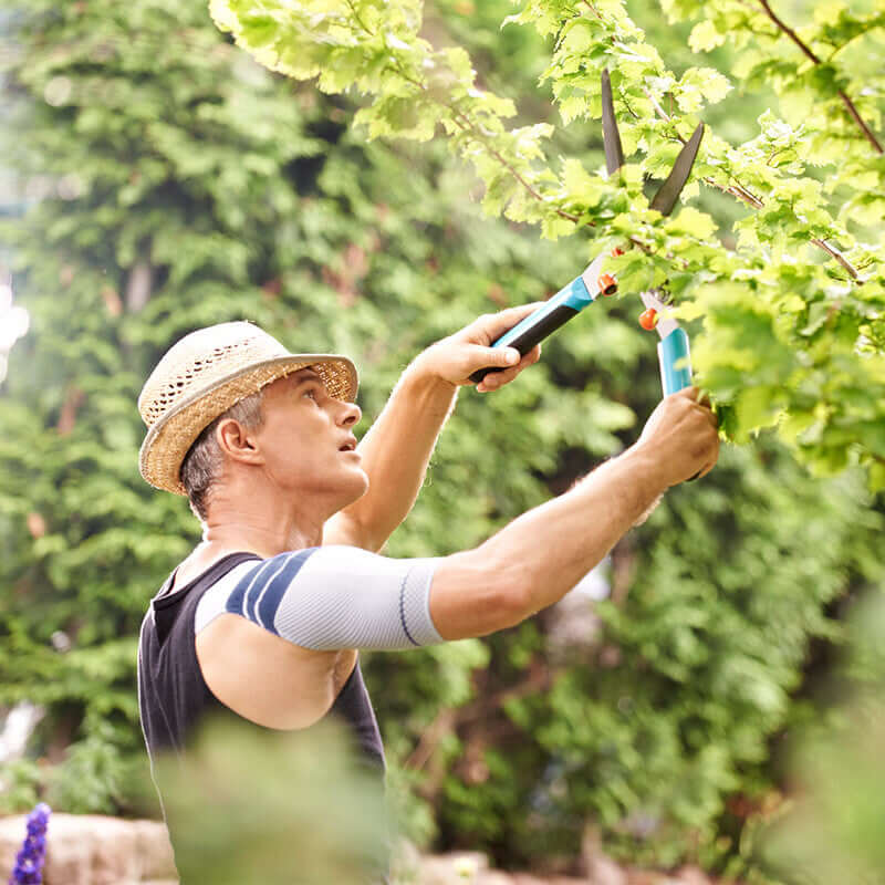 Shoulder brace worn by a man trimming shrubs with garden shears outdoors, showing active use and range of motion