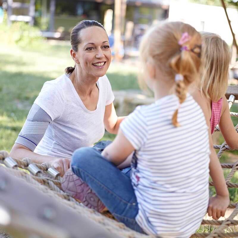 Shoulder brace worn on left arm by smiling woman sitting outdoors on rope playground equipment with two young girls