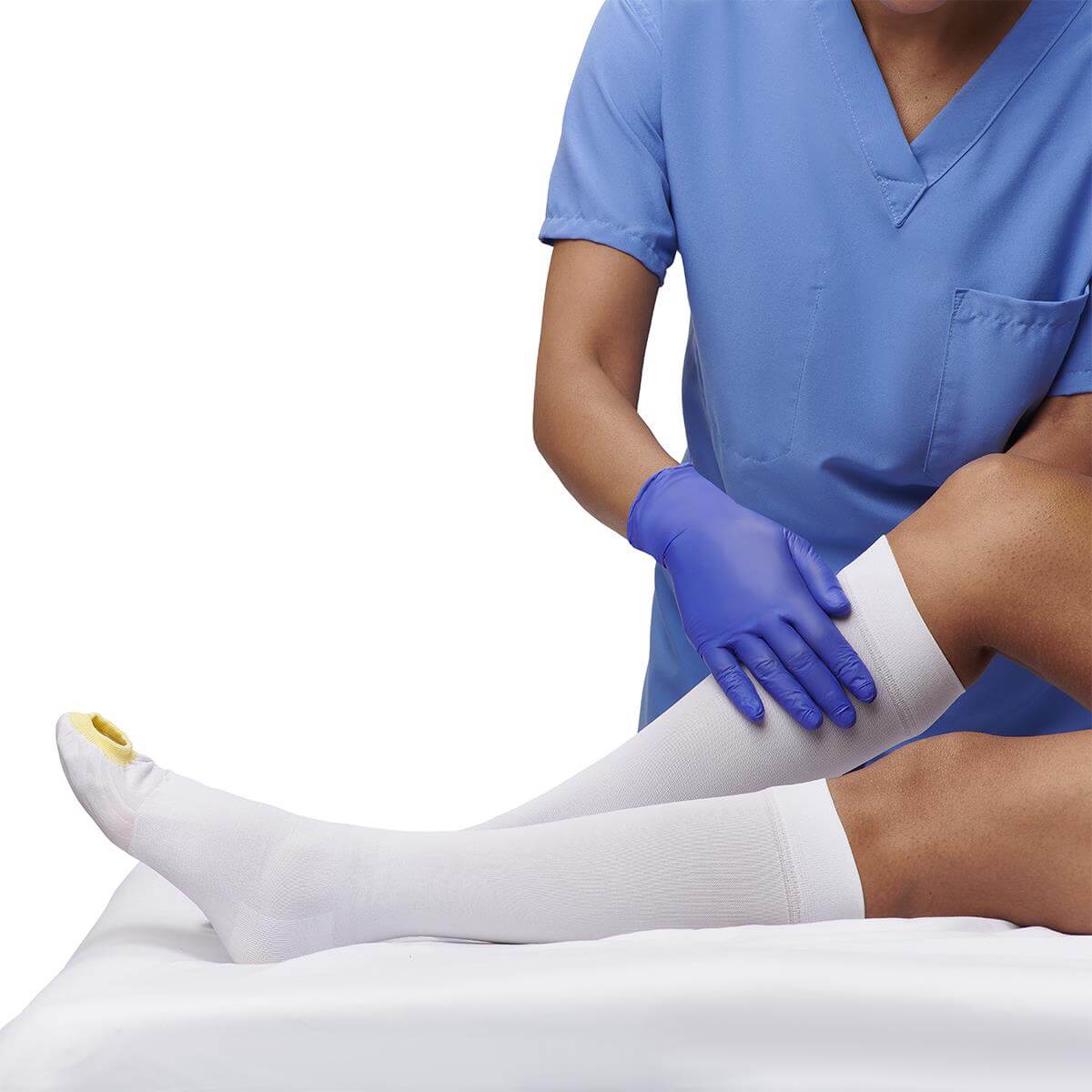 Anti-embolism stocking being applied to a patient's leg by a gloved clinician in blue scrubs, shown knee-high on leg
