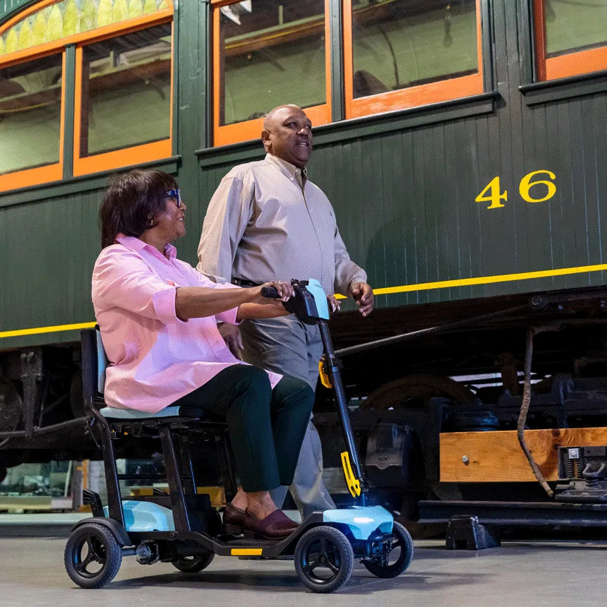 Power mobility scooter in blue ridden by a woman beside a man near a vintage train car indoors
