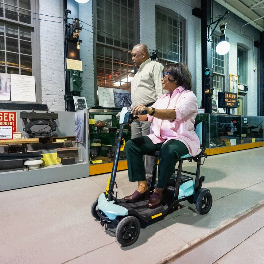 Power mobility scooter in light blue ridden by a woman indoors at a museum, with a man walking alongside her