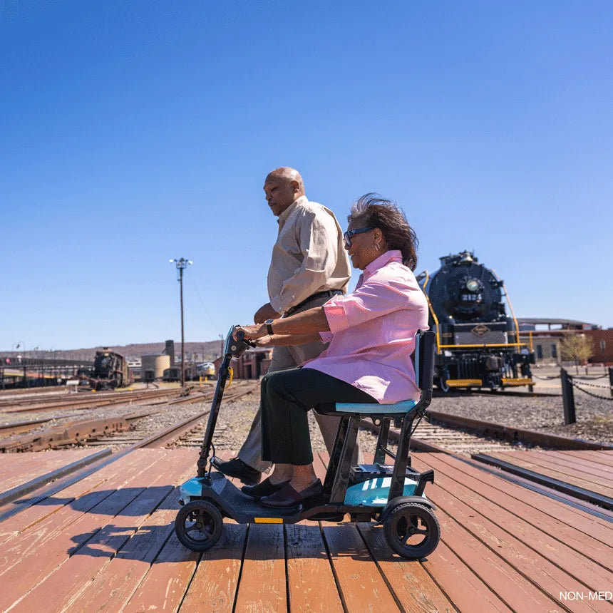 Pride Go-Go Super Portable mobility scooter in teal, ridden by a woman outdoors at a train yard on a sunny day