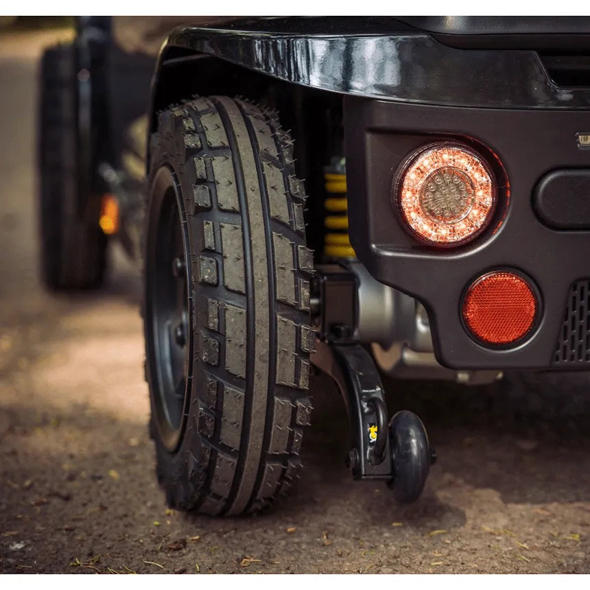 Power scooter rear wheel close-up showing rugged knobby tire tread, red LED tail light, and anti-tip wheel on black body