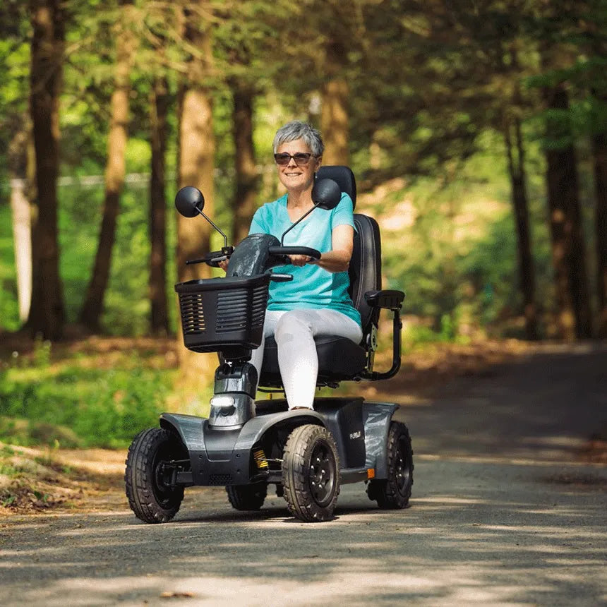 Pride Pursuit 2 power mobility scooter ridden outdoors on a tree-lined path by a smiling older woman