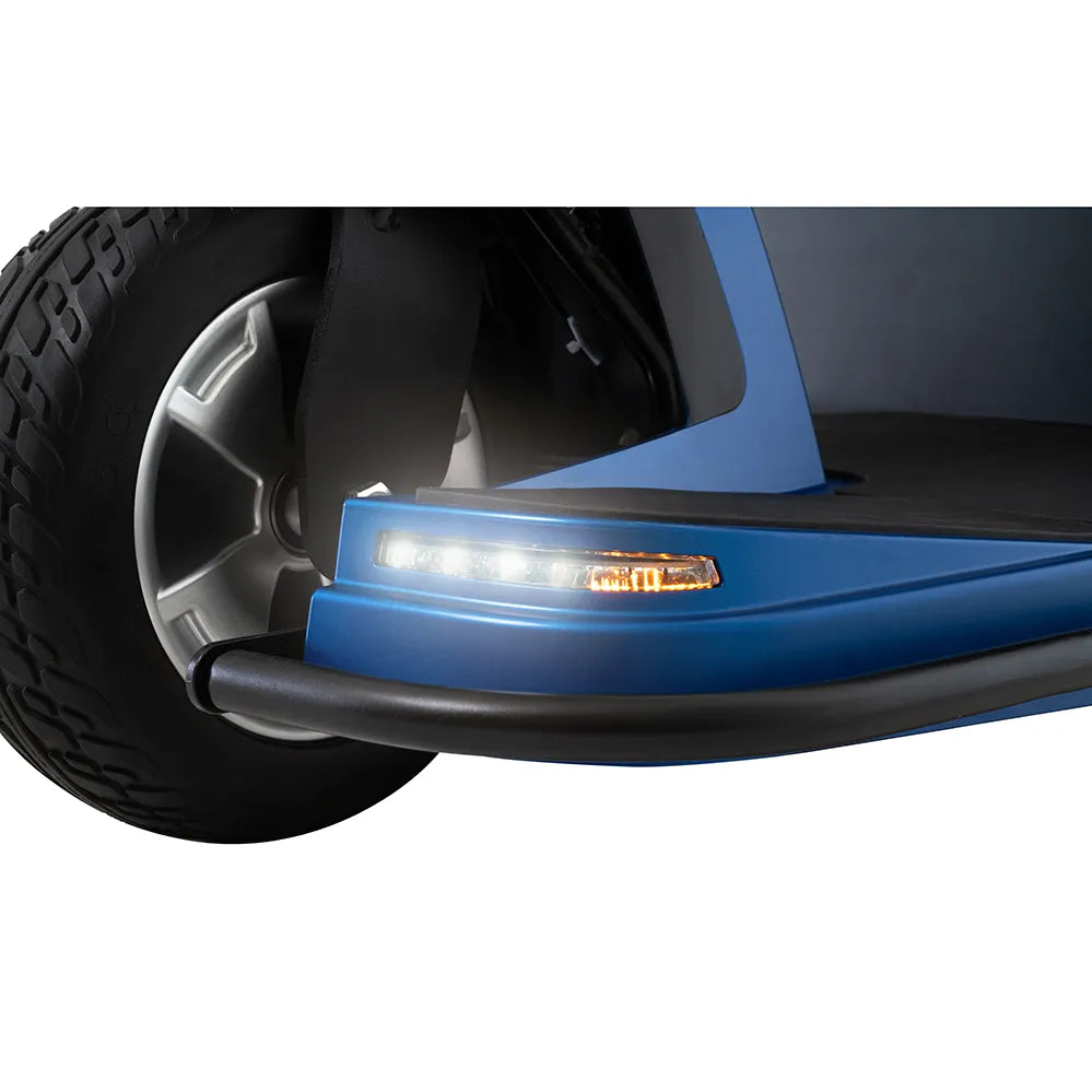 Power wheelchair front corner detail showing blue body panel with LED headlight, amber turn signal, bumper guard, and large