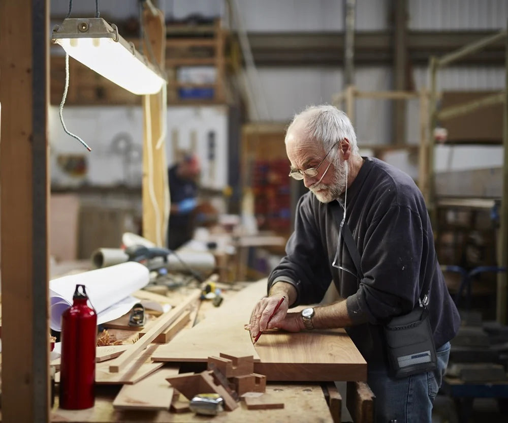 Portable oxygen concentrator user, an older man wearing a nasal cannula, marking wood at a workbench in a busy workshop