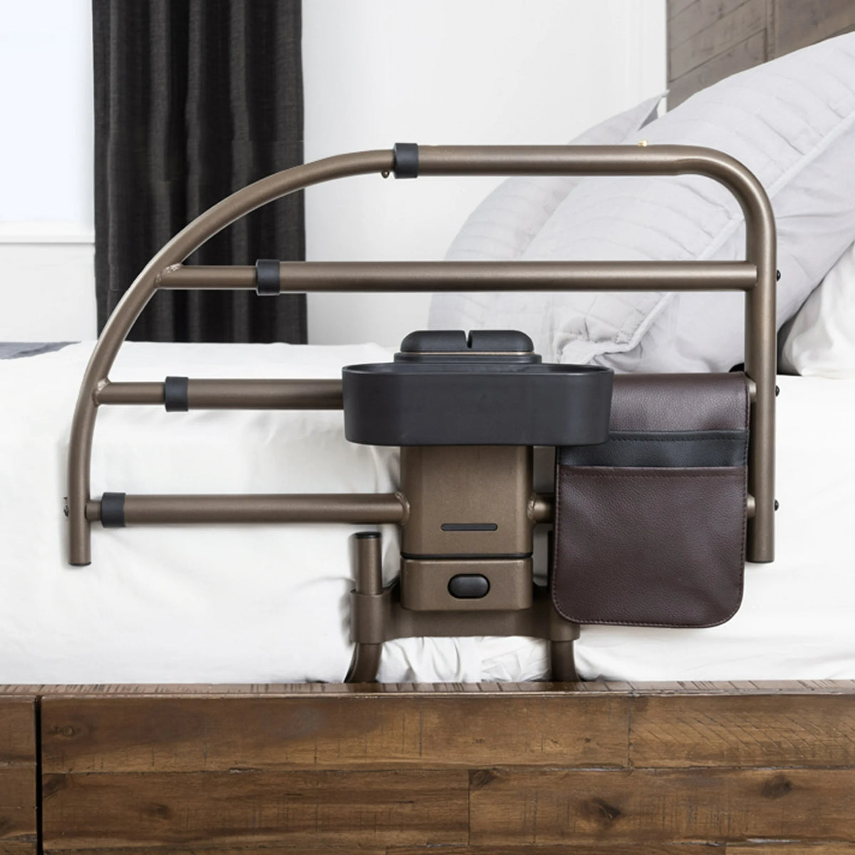 Bed rail with bronze tubular frame, attached organizer tray, and brown leather storage pouch, shown on a white-sheeted bed