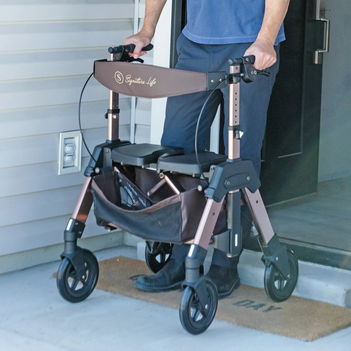 Rollator walker with seat, padded backrest, and storage bag being guided through a doorway by a person in blue clothing