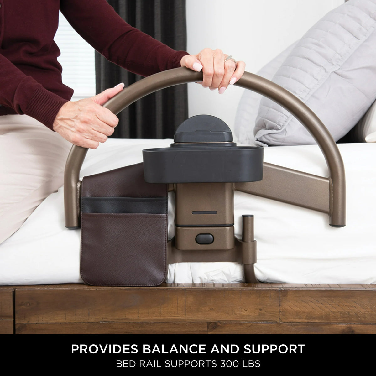 Bed rail with arched bronze handle, side pocket, and storage caddy mounted on a white bed, gripped by a person's hands