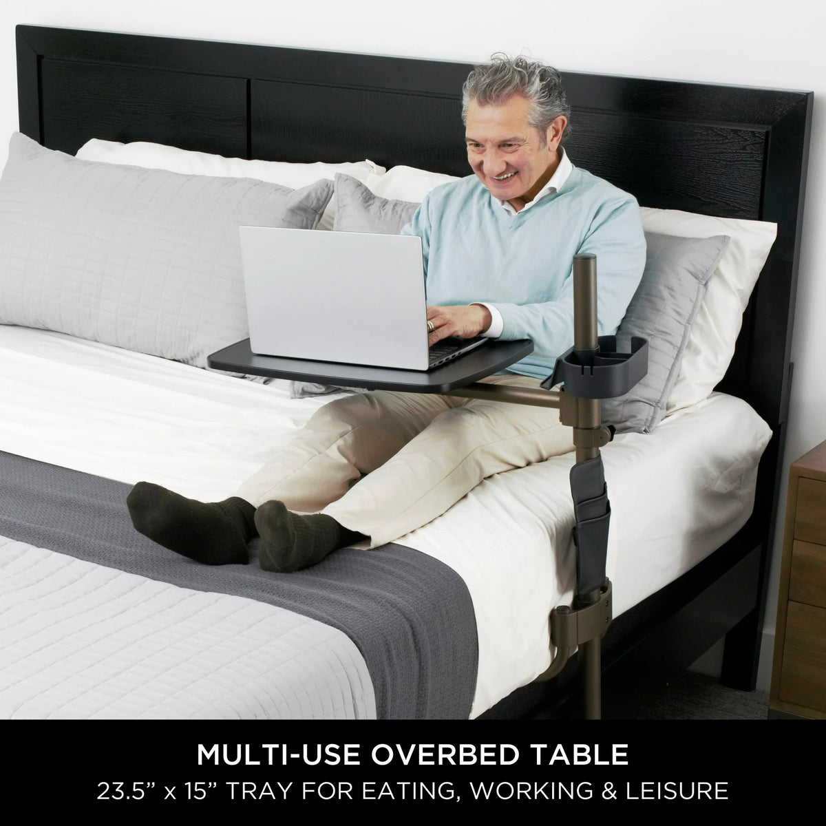 Overbed table with swivel tray and cup holder used by older man working on a laptop while sitting up in bed