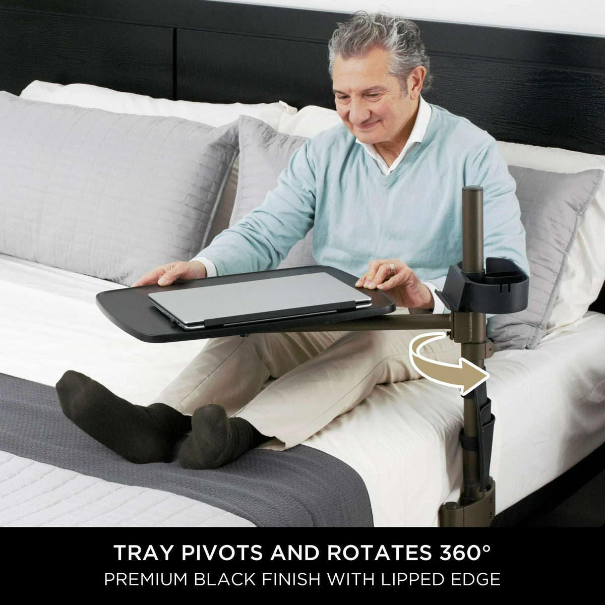 Overbed table with swivel tray and cup holder used by older man sitting in bed with a laptop, black finish with lipped edge