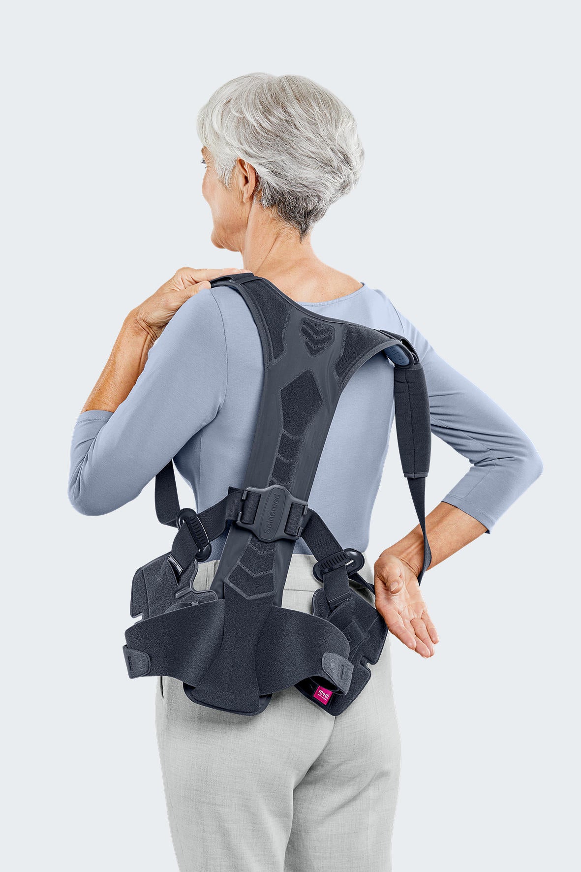 Spinomed spine brace worn by an older woman, rear view showing dark gray structured back panel with shoulder straps and