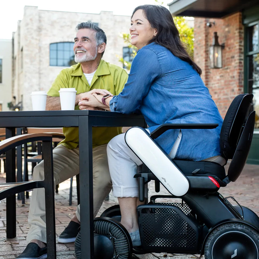 Power wheelchair user sitting at an outdoor café table with a companion, both smiling, WHILL Model C2 in black and white