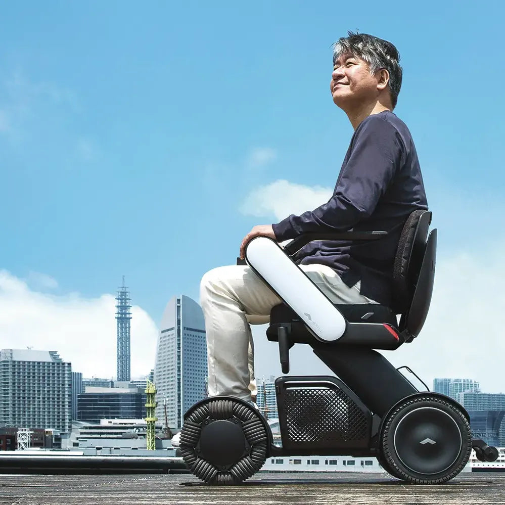 Power wheelchair with sleek black and white frame, ridden outdoors by a man against a city skyline and blue sky