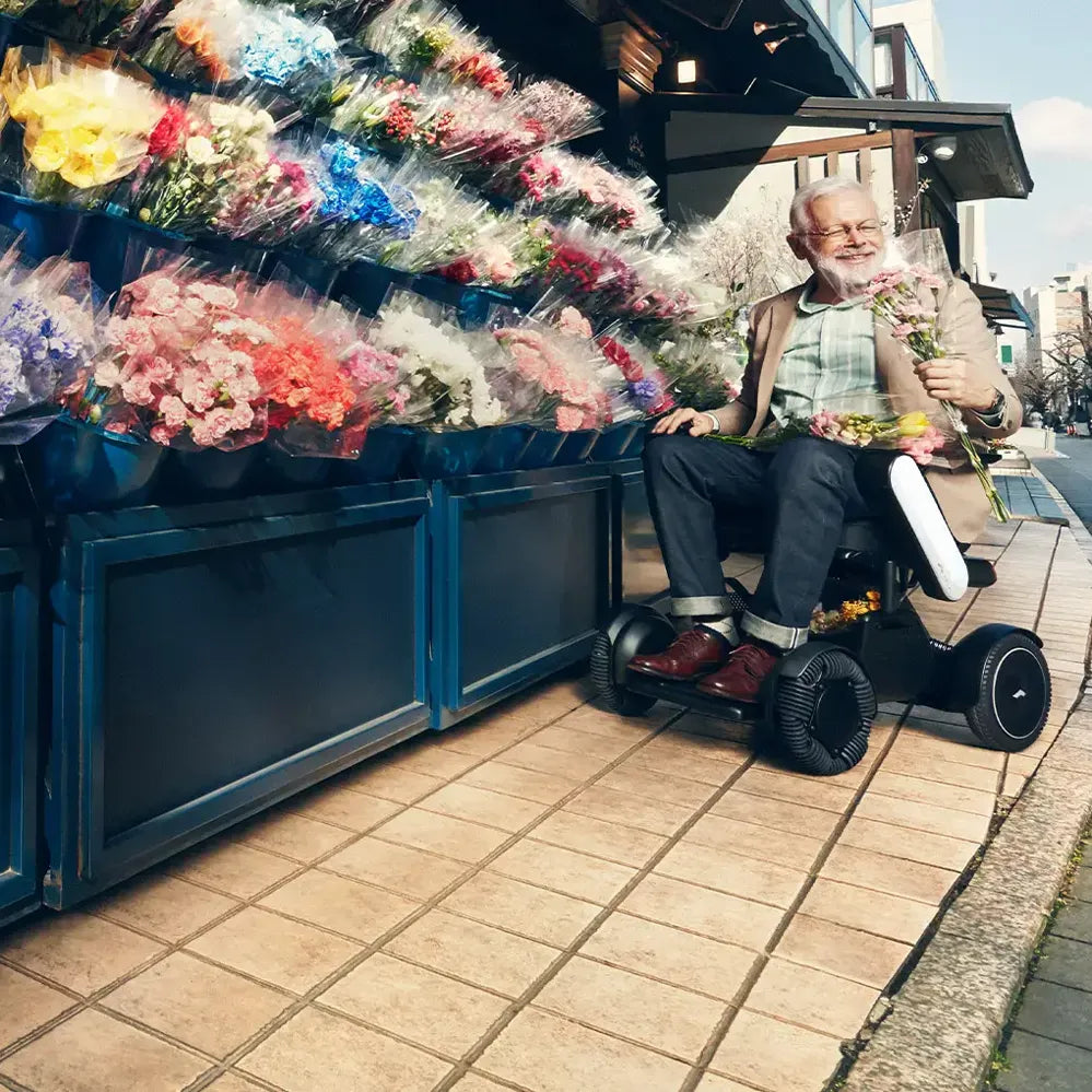 Power wheelchair with an older man smiling while browsing a colorful outdoor flower market on a city sidewalk