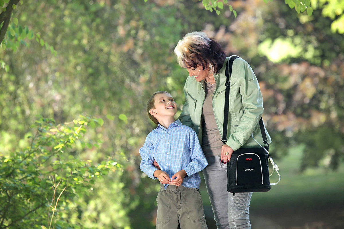 Portable oxygen concentrator carry bag worn over shoulder by woman walking outdoors with a young boy in a park setting