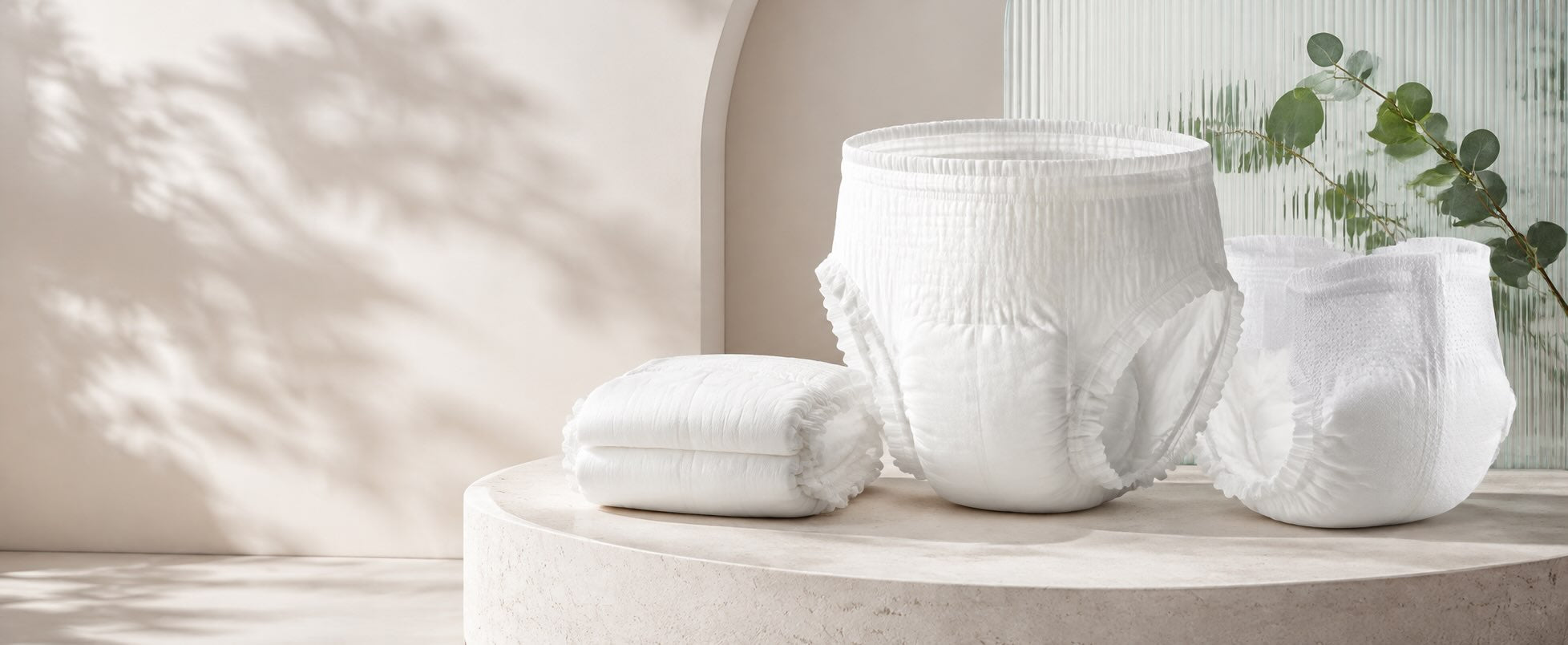 Three white disposable diapers arranged on a round stone pedestal, with a matching folded diaper beside them. The setting is a softly lit interior with warm beige walls, leaf shadows, and a small green plant in the background.