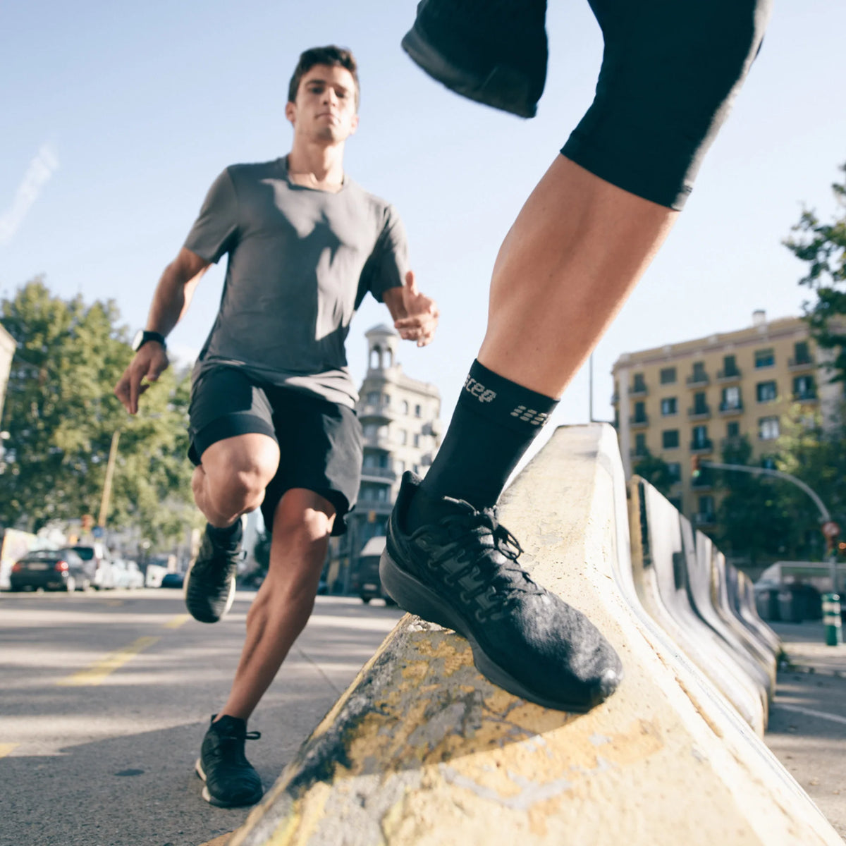 Compression ankle sleeve worn by two runners on an urban street, shown in black from a low ground-level angle