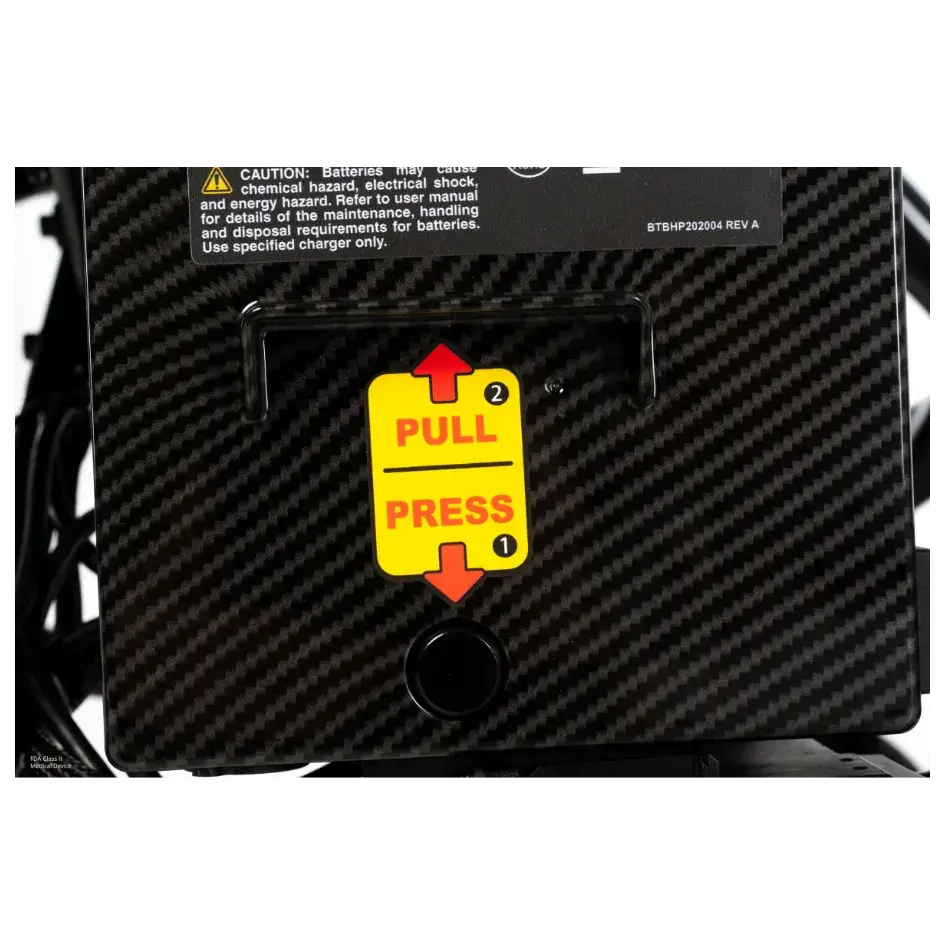 Power wheelchair battery compartment close-up with yellow Pull/Press release label on carbon fiber panel