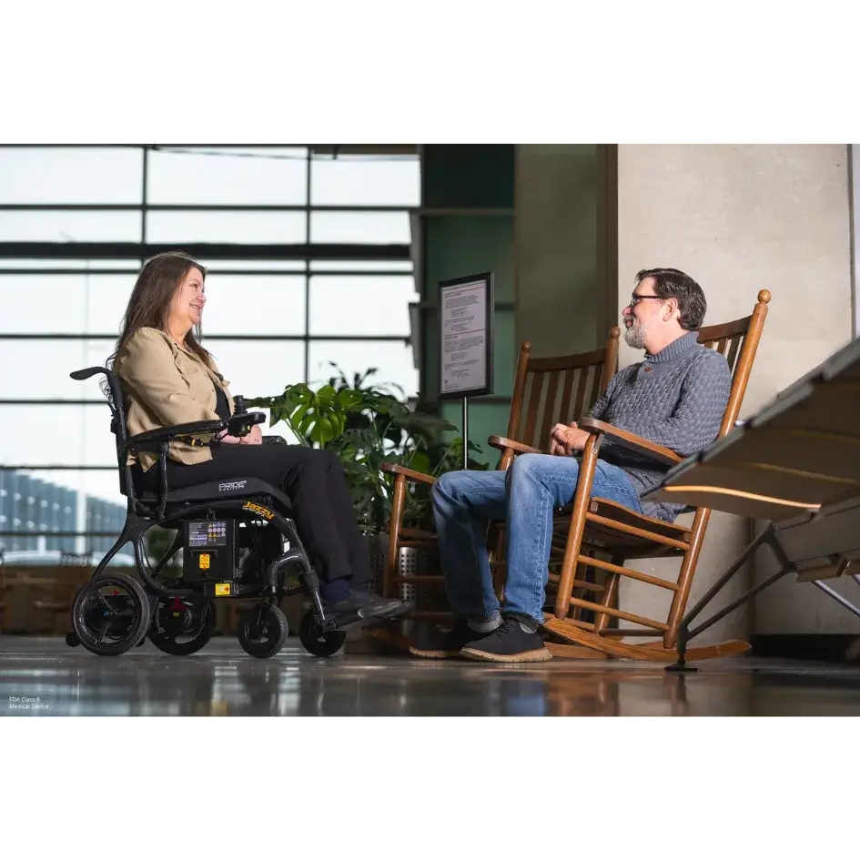 Power wheelchair with black frame and joystick control, shown with a woman seated in it conversing with a man in a rocking