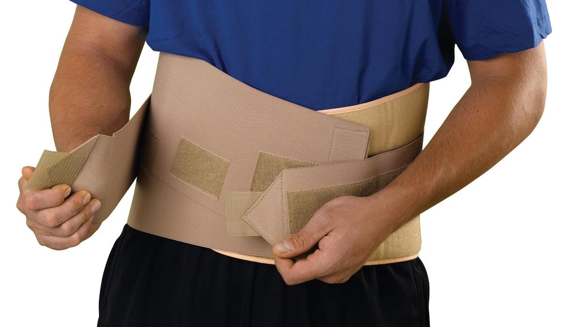 Lumbar back support brace in beige worn by a man, shown being fastened with hook-and-loop straps around the lower back