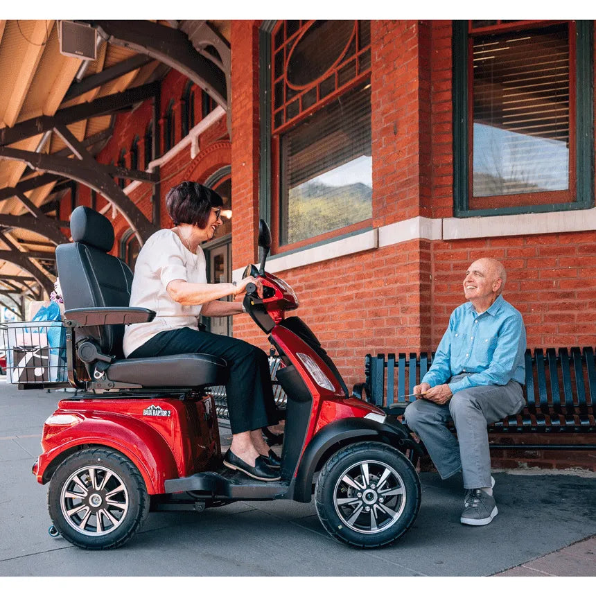 Pride Mobility Baja Raptor 2 four-wheel mobility scooter in red, ridden by a woman conversing with a man on a bench outdoors