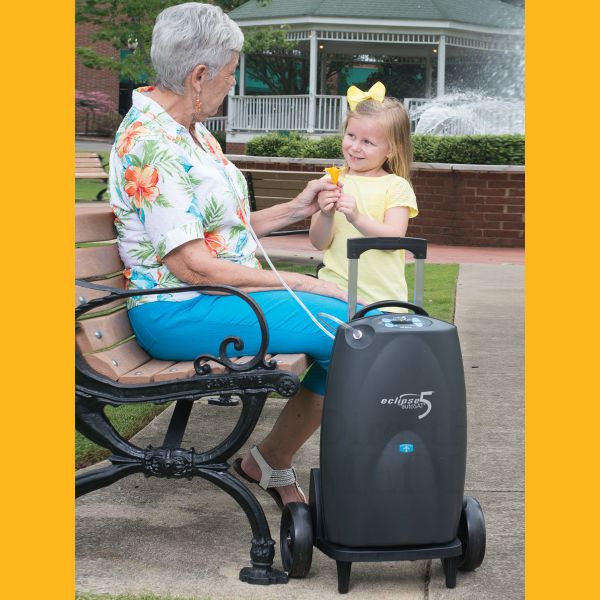 Eclipse 5 portable oxygen concentrator on wheels beside an older woman seated on a park bench with a young girl nearby