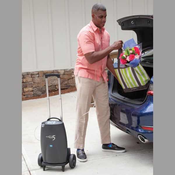Portable oxygen concentrator on wheeled cart beside man unloading flowers from a car trunk outdoors