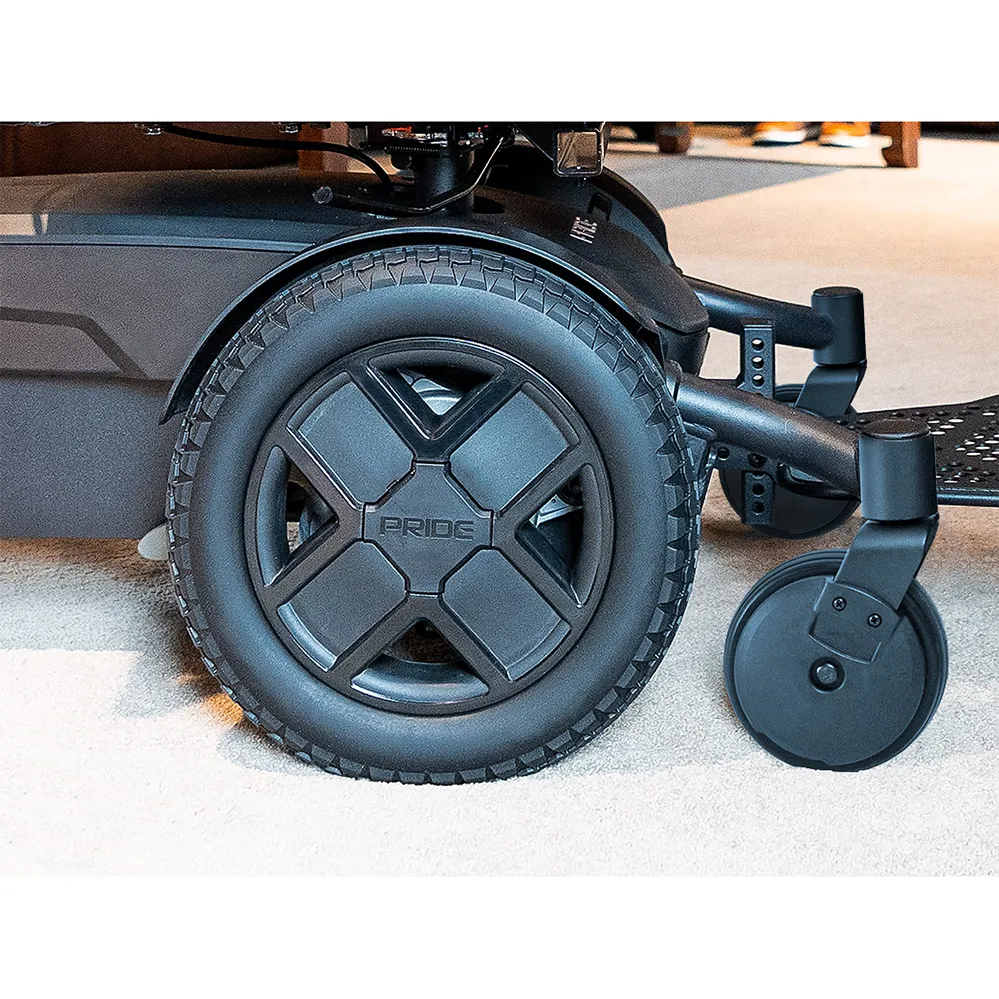 Power wheelchair close-up showing large rear drive wheel with treaded tire and smaller front caster wheel on carpet flooring