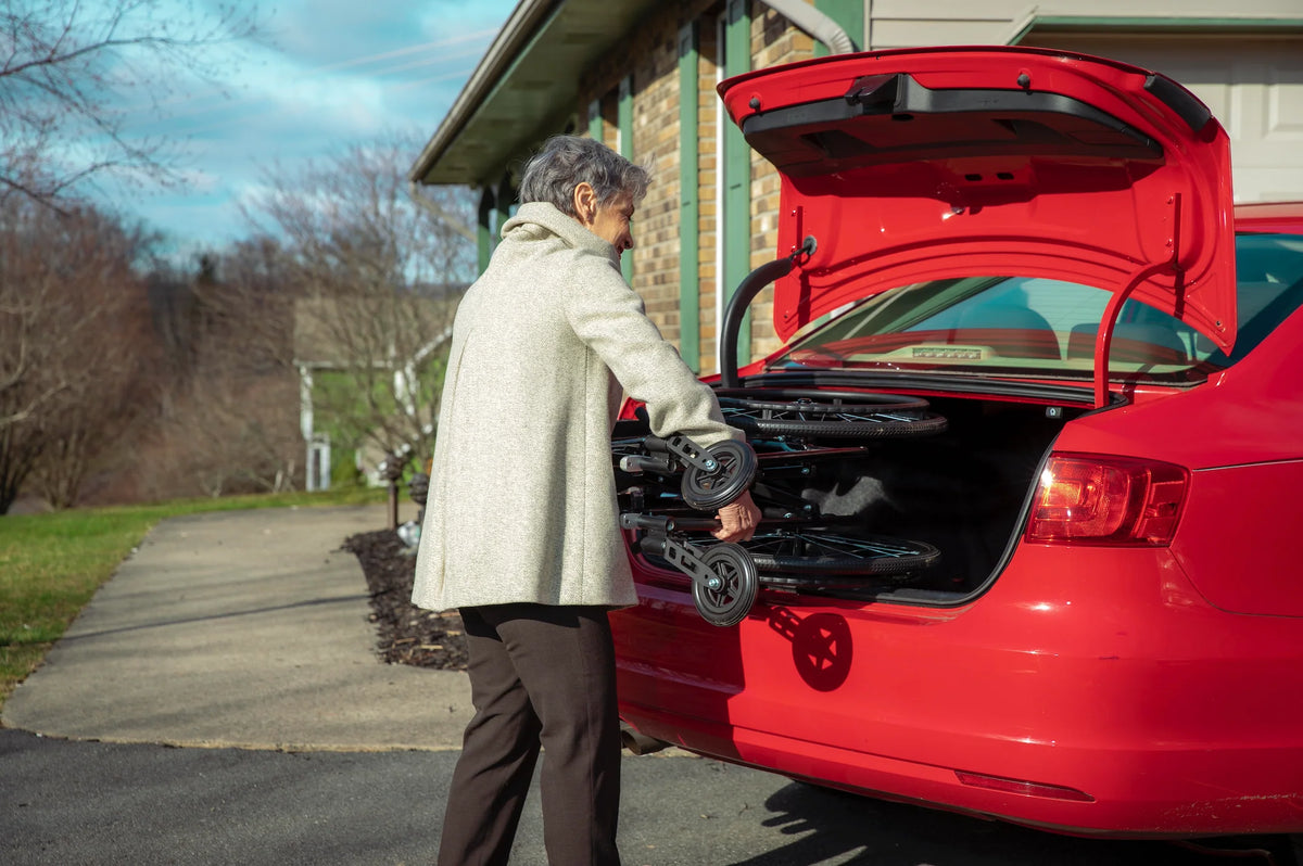 Lightweight wheelchair being loaded into a red car trunk by an elderly woman in a residential driveway