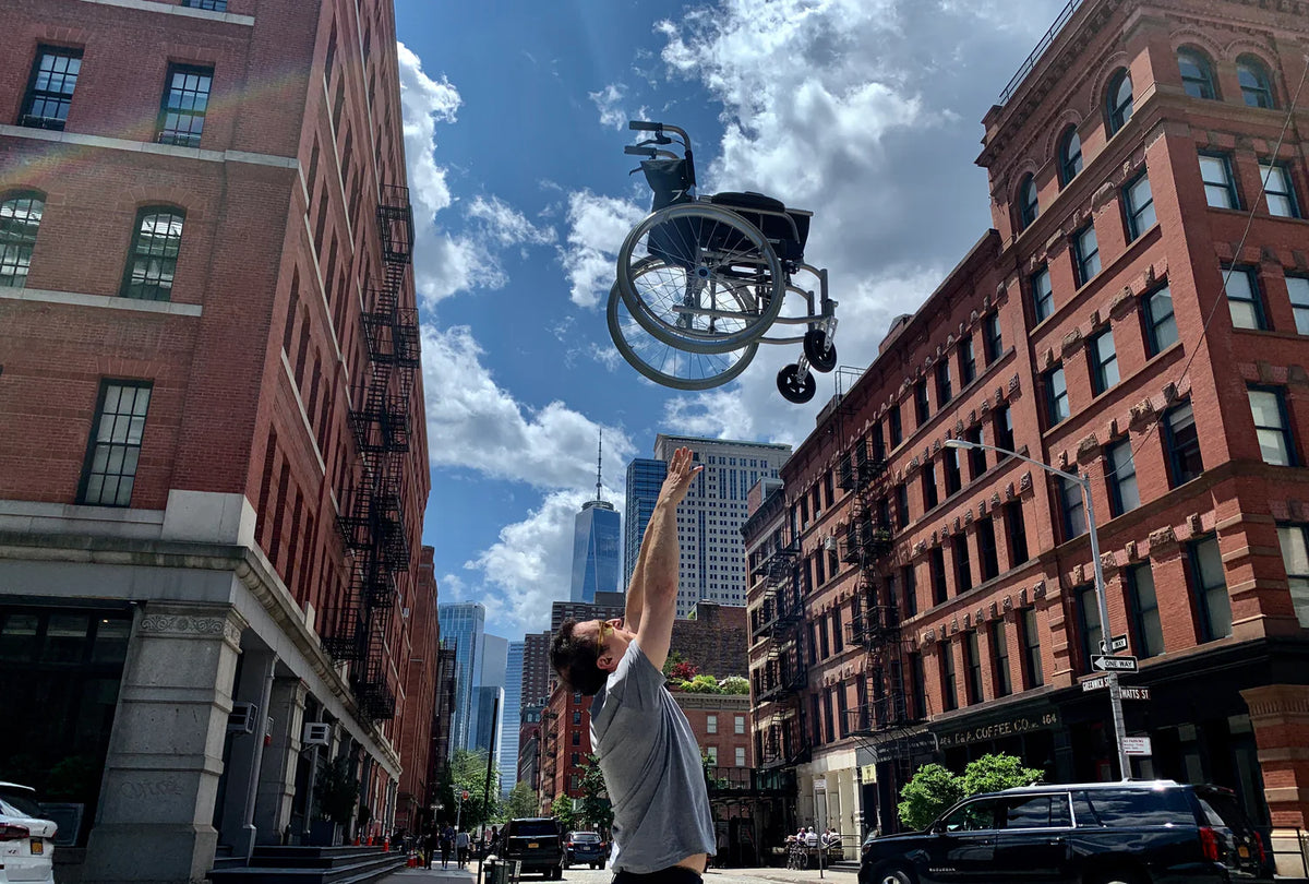 Lightweight wheelchair tossed in the air by a man on a city street, with urban buildings and blue sky in the background