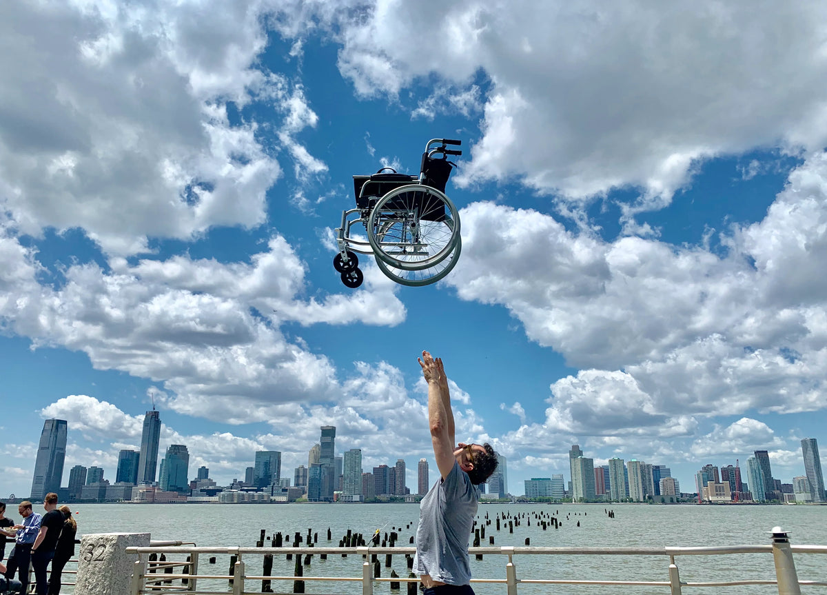 Lightweight wheelchair tossed into the air by a man on a waterfront boardwalk with a city skyline in the background