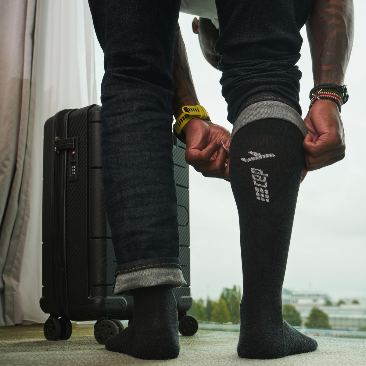 Compression flight socks in black being pulled on by a person standing next to a carry-on suitcase near a window