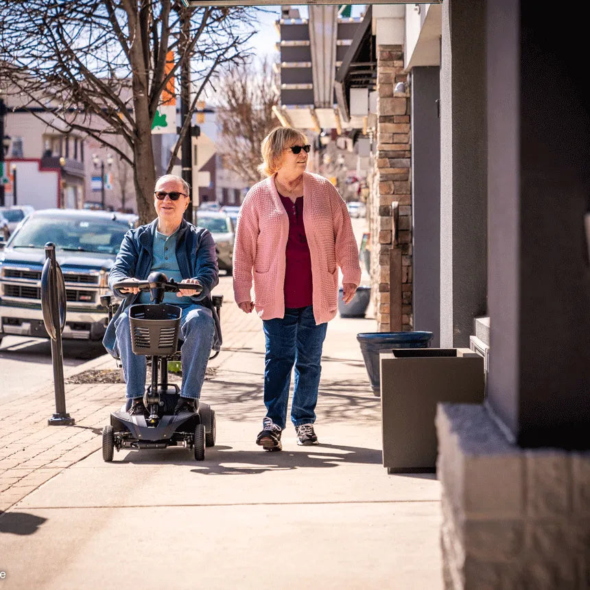 Power mobility scooter ridden by a man alongside a woman walking on a sunny downtown sidewalk