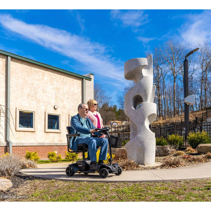 Power mobility scooter ridden by a man outdoors on a sunny day, with a woman walking alongside near a stone sculpture