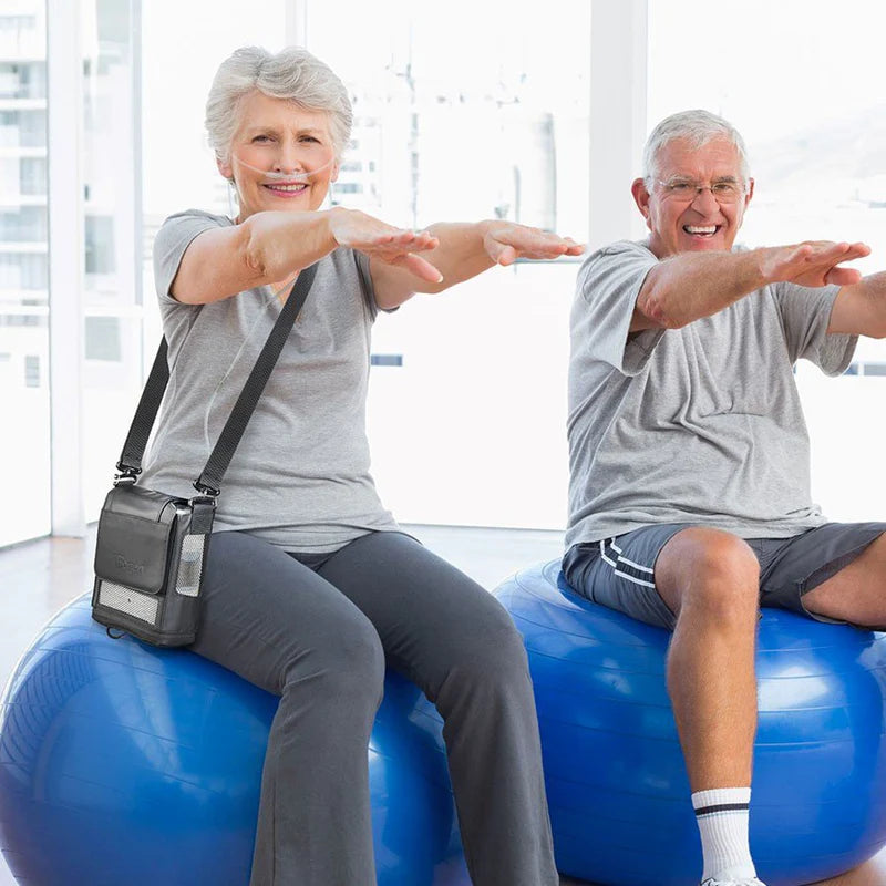 Portable oxygen concentrator worn crossbody by a smiling senior woman exercising on a blue balance ball with a male companion