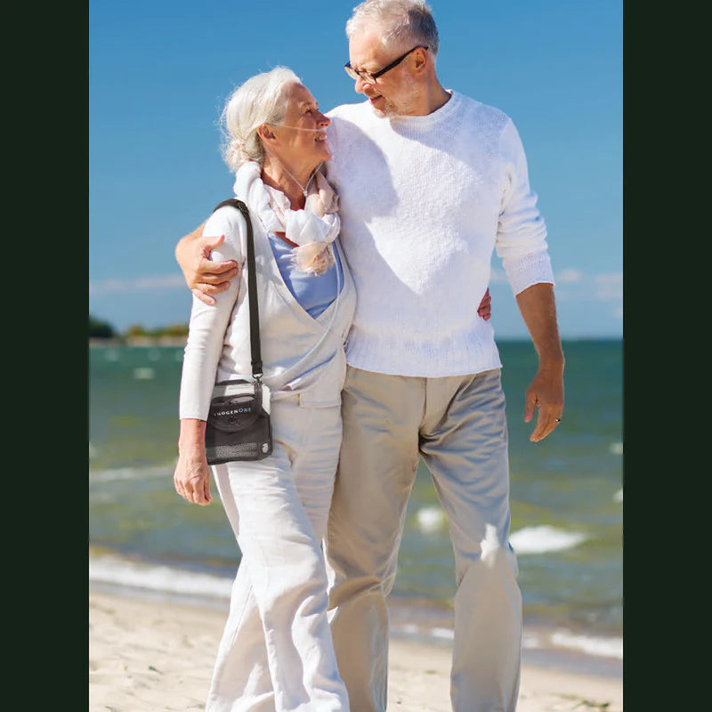 Portable oxygen concentrator worn via shoulder bag by older woman walking on beach with male companion on sunny day