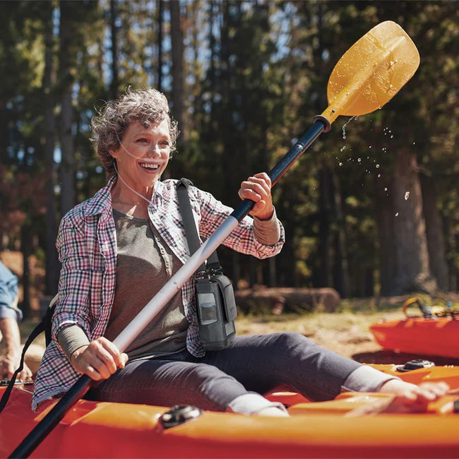 Portable oxygen concentrator worn by smiling older woman kayaking on a lake surrounded by trees