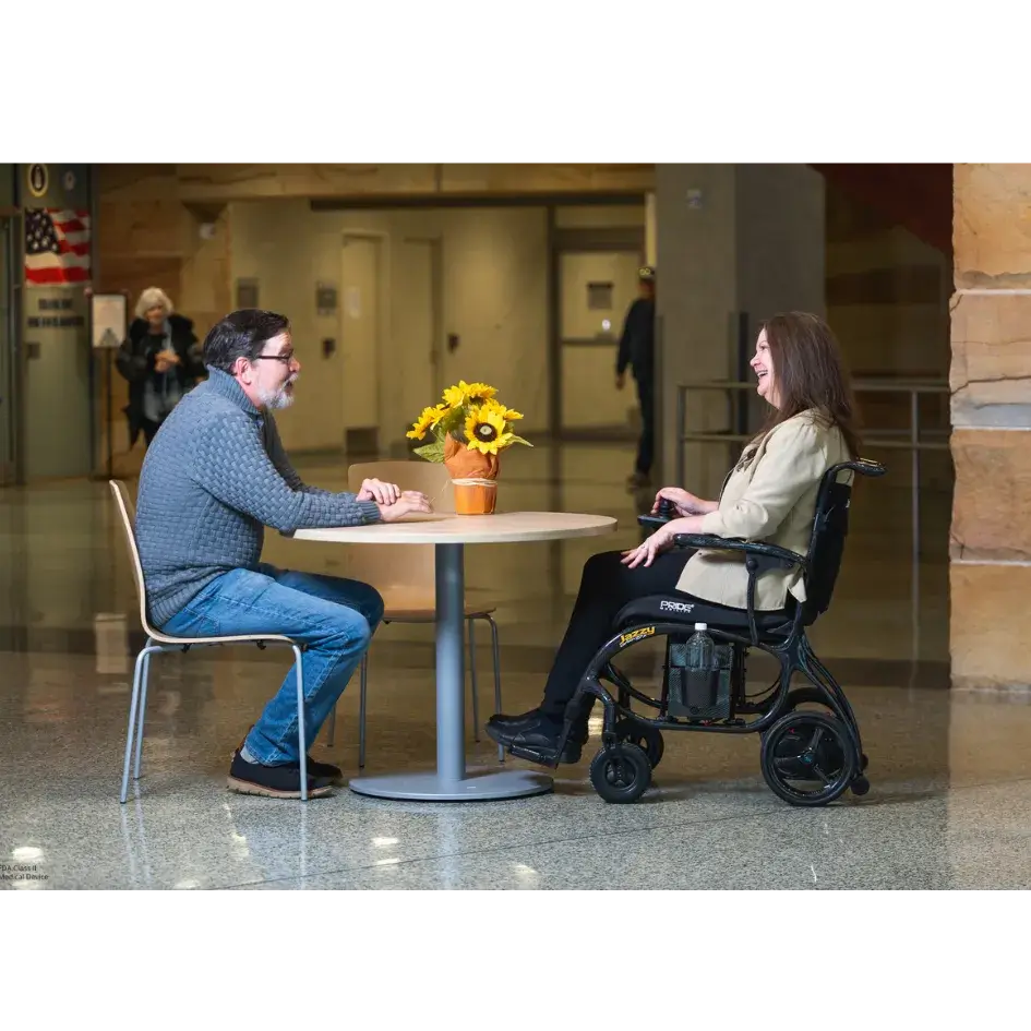 Power wheelchair user seated at a round café table conversing with a companion indoors, Jazzy Carbon in black frame visible
