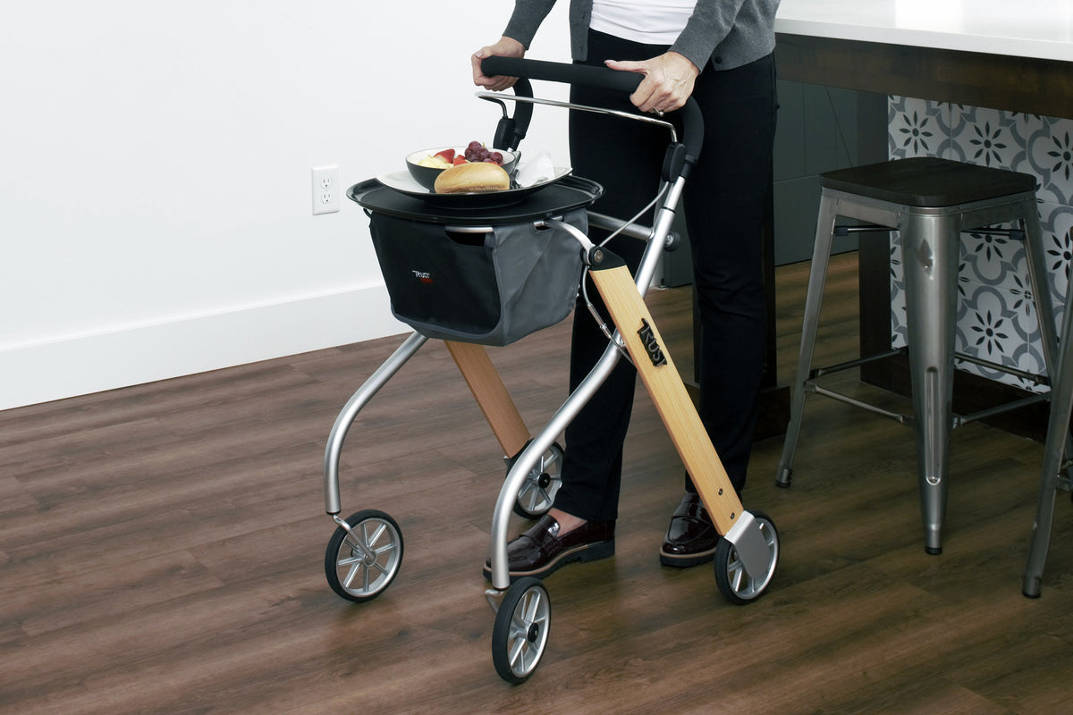 Indoor rollator with wood-accent frame, black basket, and tray holding food, used by a person walking on hardwood floors