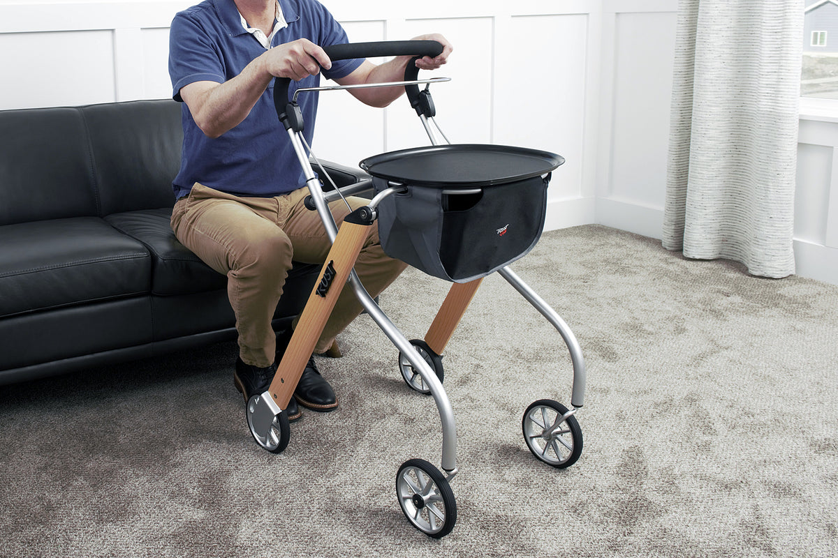Indoor rollator with wood-tone frame, padded handles, black tray, and basket bag, held by a seated man near a sofa