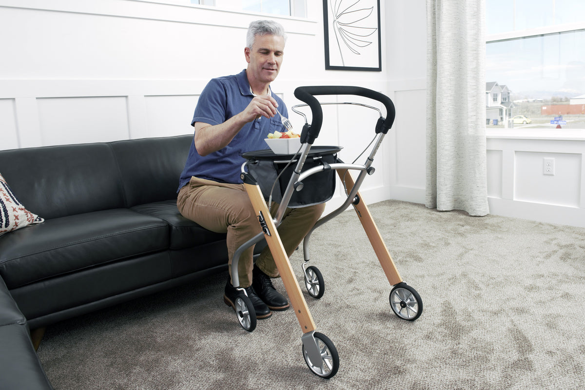 Indoor rollator with wood-tone frame, tray, and storage bag beside a man seated on a sofa eating from a bowl
